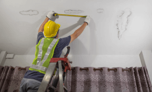 Man in a hard hat on a ladder measuring the size of a roof leak stain on the ceiling with a tape measurer.